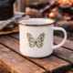 Butterfly enamel mug on wooden table near campfire, lightweight camping coffee cup for outdoor and travel use