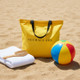 Yellow beach tote bag on sand beside towel and beach ball in a summer setting
