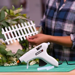  Person using a white Arrow hot glue gun while assembling a small decorative white picket fence craft on a worktable.