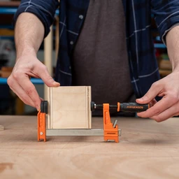 Person tightening an orange Pony Jorgensen bar clamp to secure a small wooden box-shaped workpiece on a workbench.