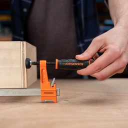 Person tightening the black handle of an orange Pony Jorgensen bar clamp to secure a wooden workpiece on a workbench.