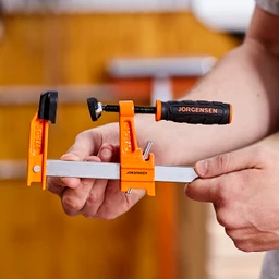 Person holding an orange Pony Jorgensen bar clamp with a black comfort-grip handle and silver steel bar in a workshop.
