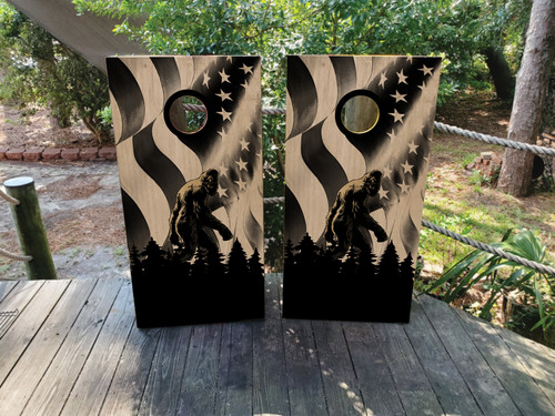 Pair of cornhole boards featuring a black and white American flag background with a silhouette of Bigfoot walking through a pine forest, creating a dramatic, high-contrast wilderness scene.