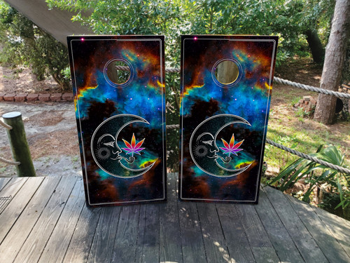 Pair of cornhole boards with a vibrant galaxy background featuring a crescent moon, an Om symbol, and a colorful cannabis leaf, all outlined in white with glowing cosmic accents.