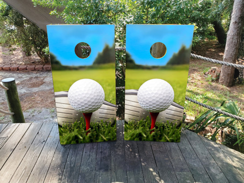Pair of cornhole boards featuring a close-up of a white golf ball sitting on a red tee, set against a bright green fairway and blue sky with trees in the distance, creating a sunny golf course scene.