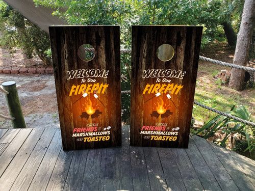 "Welcome to Our Firepit" cornhole boards with campfire design, marshmallows on sticks, and rustic wood background, placed outdoors on a wooden deck surrounded by greenery.