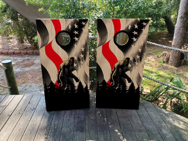 Pair of cornhole boards with a stylized American flag background in black, white, and red, featuring a detailed Bigfoot silhouette walking through a pine forest.