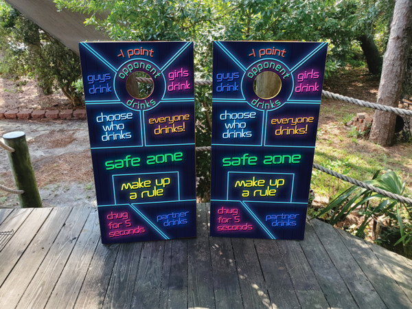 Cornhole boards featuring different phrases of a drinking game in neon colors. Phrases include safe zone, guys drink, girls drink, opponent drinks, choose who drinks, everyone drinks, shotgun, safe zone, toast, chug for 10 seconds, partner drinks, make up a rule