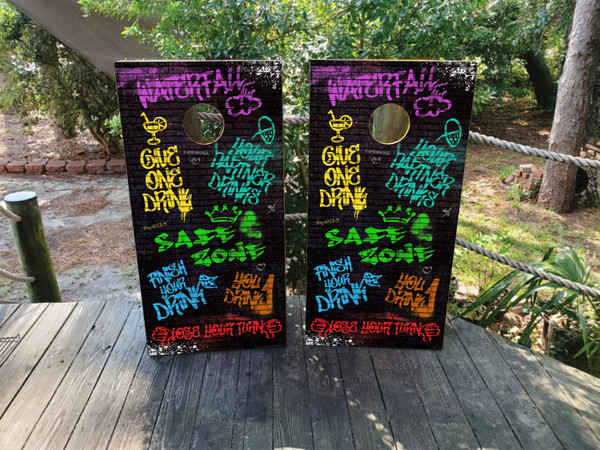 Cornhole boards featuring different phrases of a drinking game in neon colors. Phrases include safe zone, senors drink, senoritas drink, you drink, choose who drinks, everyone drinks, shotgun, safe zone, toast, chug for 10 seconds, partner drinks, everyone drinks, jumping  jacks