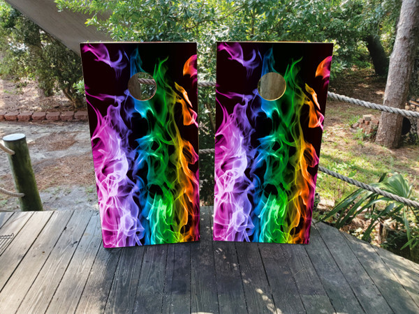 Pair of cornhole boards with vibrant rainbow-colored flames rising from the bottom against a black background, blending colors like pink, purple, blue, green, and yellow in a fiery pattern.