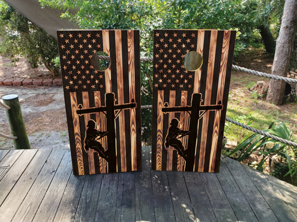 Two wooden boards featuring a rustic American flag design with a silhouette of a lineman climbing a utility pole in front of a large cross, symbolizing faith, strength, and dedication.