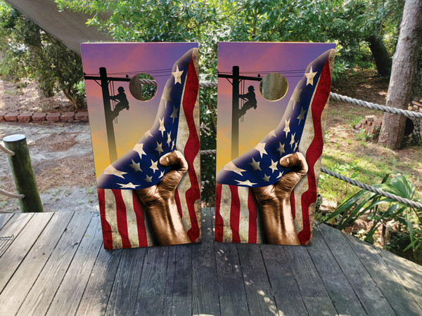 Cornhole boards featuring a hand pulling back a USA flag and revealing a silhouette of a lineman climbing a pole