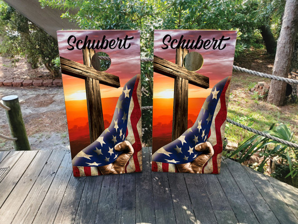 Cornhole boards featuring a hand pulling back a USA Flag to reveal a sunset and cross over a mountain landscape
