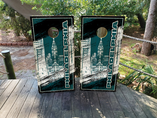 Pair of cornhole boards featuring a white Philadelphia skyline illustration with a distressed grunge overlay, set against a dark green and black background with bold “PHILADELPHIA” text running vertically along the side.