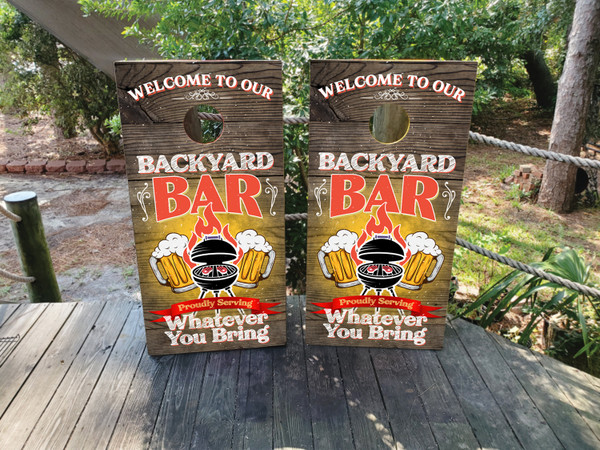 Two cornhole wraps with a rustic wood design featuring the text 'Welcome to Our Backyard Bar,' a flaming grill, beer mugs, and the phrase 'Proudly Serving Whatever You Bring,' perfect for barbecue and outdoor party themes