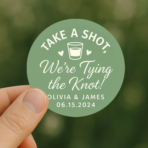 Hand holding a small wedding favor bottle featuring a sage green “Take a Shot” label, highlighting the design size and glossy finish for product scale reference.