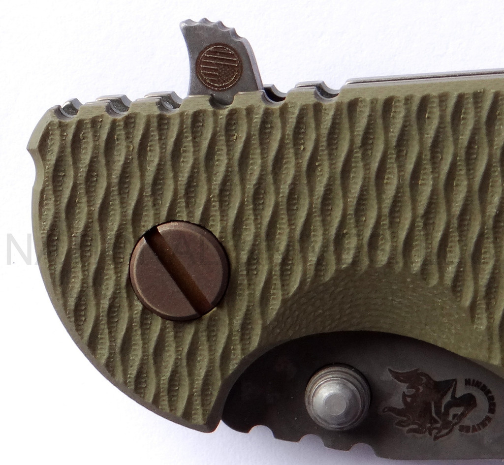 Rick Hinderer Knives Battlefield Pick Up XM-18 3.5" Harpoon Spanto folding knife with thumb stud and flipper tab - close up of presentation side pivot shown.  M390 stainless steel plain edge blade.  OD green G-10 handle with titanium lock side.  Model K25520SP00.
For reference only.