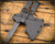 RMJ Tactical 3V Syndicate Raven tomahawk - shown close up of head in sheath obverse.  Smoke Gray Cerakote CPM-3V steel plain edge head.  Dirty Olive G-10 handle.
For reference only.