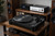 Audio-Technica AT-LP7X turntable in matte black on a walnut wood hi-fi rack with a vinyl record spinning, flanked by floor-standing speakers and integrated amplifier in a warm ambient listening room setting