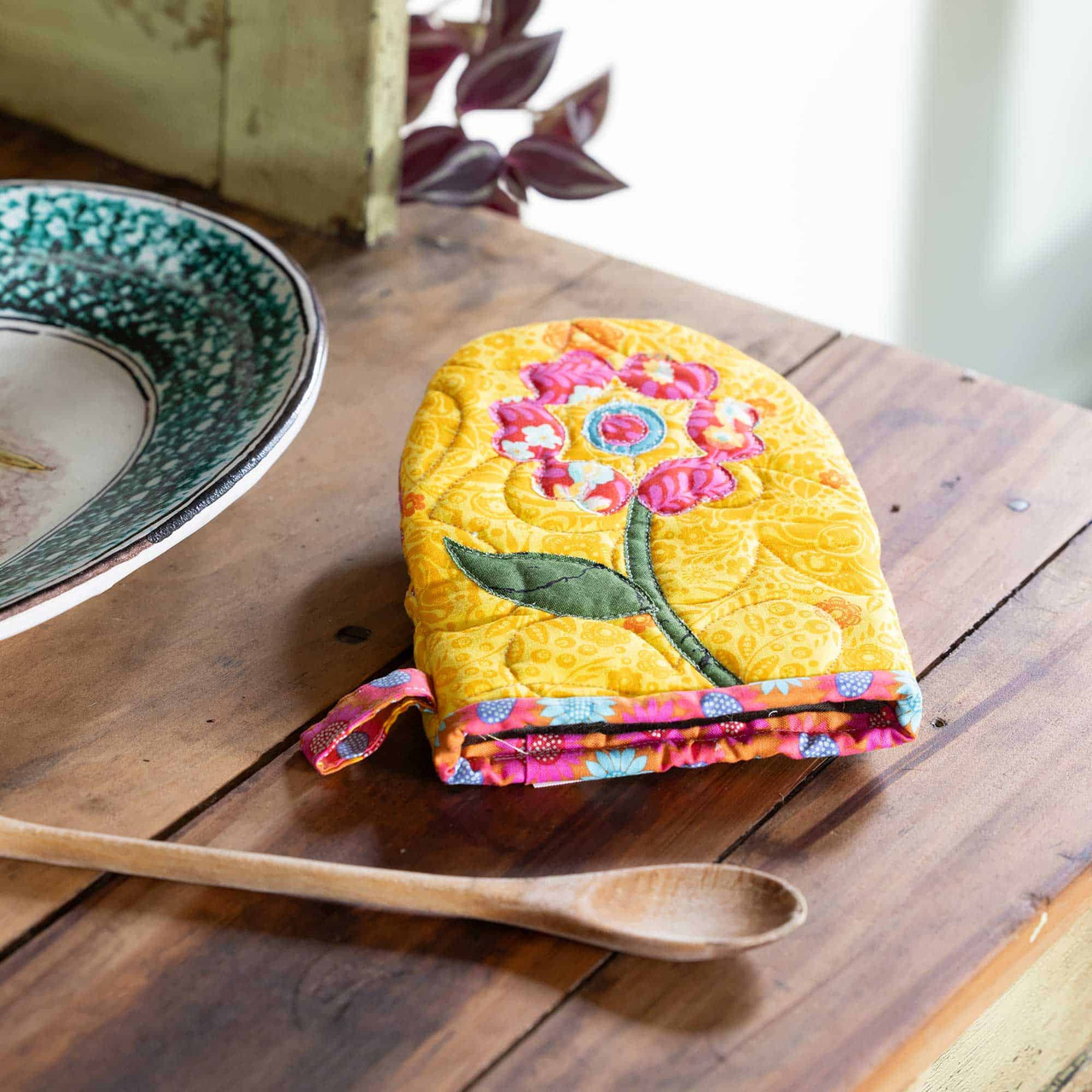 Bright floral oven mitt on a wooden kitchen table with a wooden spoon nearby.