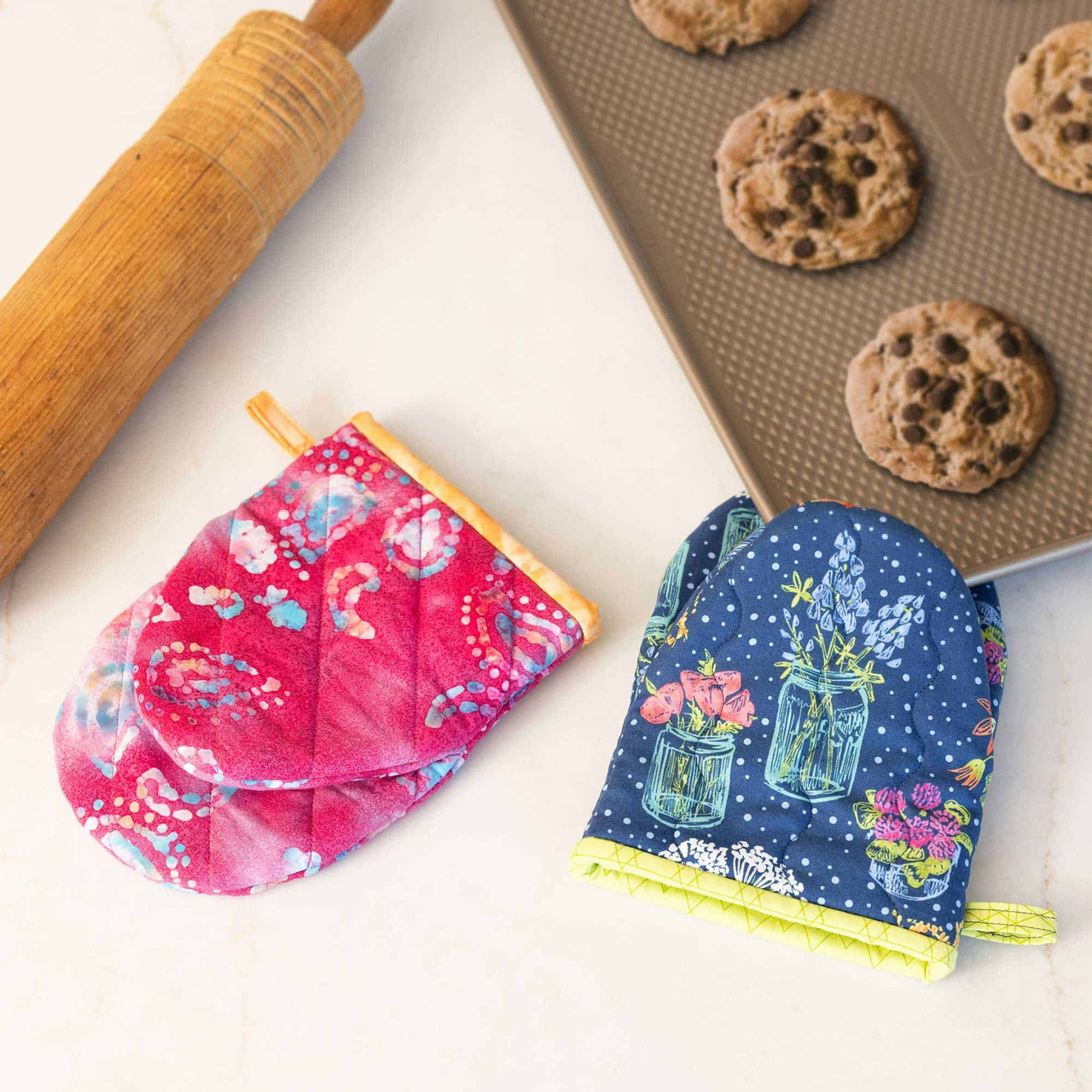 Colorful oven mitts with floral patterns next to cookies and a rolling pin.