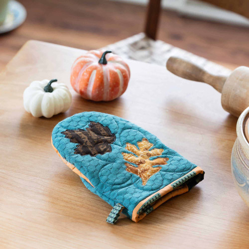 A quilted oven mitt with leaf designs beside small decorative pumpkins.