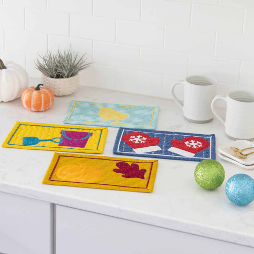 Quilted mug rugs with seasonal designs displayed on a white kitchen countertop.