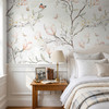 Magnolia Chinoiserie Wallpaper styled in a bright bedroom with white bedding, wood nightstand, butterflies, and soft blush magnolia branch pattern