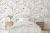 Magnolia Chinoiserie Wallpaper styled in a soft bedroom with white bedding, wood nightstand, blush magnolia flowers, and delicate chinoiserie floral walls