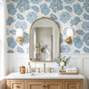 Linnea blue watercolor floral wallpaper in 26 inch repeat styled in a bathroom with gold arch mirror, brass sconces, white wainscoting, and wood vanity
