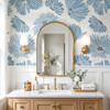 Linnea blue watercolor floral wallpaper in 52 inch repeat styled in a bathroom with gold arch mirror, brass sconces, white wainscoting, and wood vanity