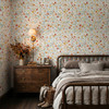 Madison wallpaper with a fine pink and yellow wildflower pattern wrapping a cottage bedroom with a dark metal bed, wooden dresser and glowing wall sconce.