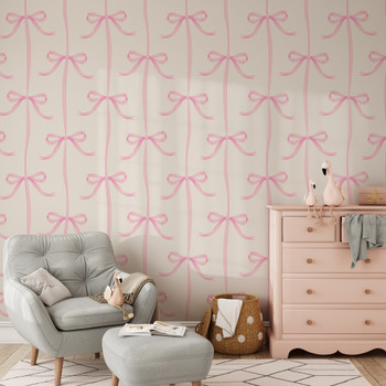 Pink bow wallpaper with large ribbon pattern in a neutral girl’s nursery with gray chair and blush dresser