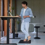 ofinto Ergonomische Stehmatte Schwarz am höhenverstellbaren Schreibtisch Elevate schwarz - Mann in hellblauem Outfit arbeitet stehend am Stehschreibtisch auf Anti-Ermüdungsmatte in modernem Büro mit Betonwand und Sitz-Stehhocker Move
