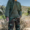 Man standing in the wood with his left hand in his pocket wearing the Olive BDU jacket shirt