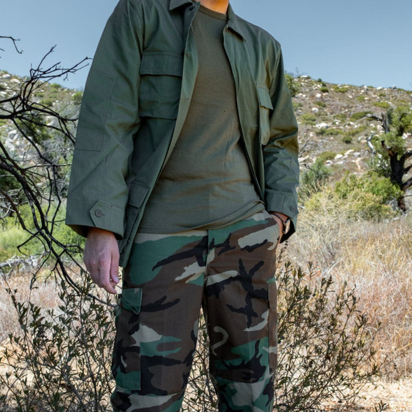 Man standing in the wood with his left hand in his pocket wearing the Olive BDU jacket shirt Man standing in the wood with his left hand in his pocket wearing the Olive BDU jacket shirt