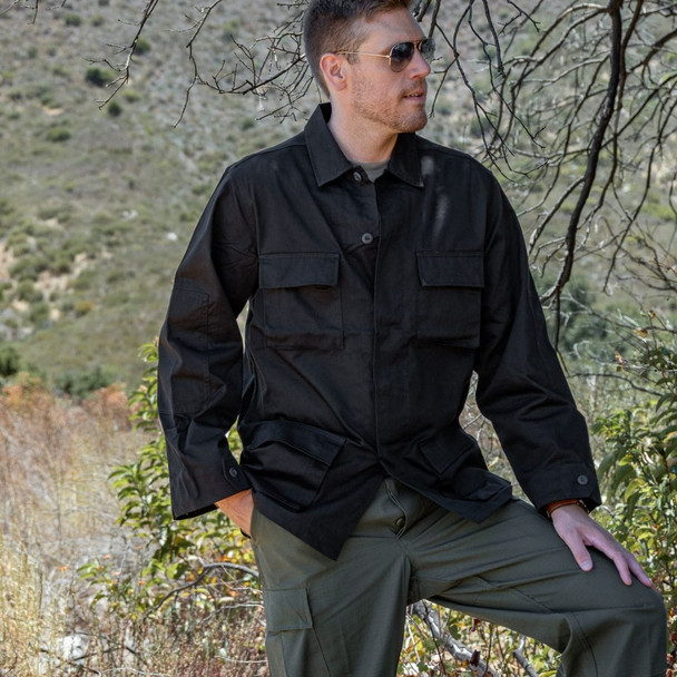 Man standing in the wood with his hand on his left leg that's propped up wearing the black BDU jacket shirt