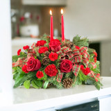 A christmas centerpiece with two red candles, roses and carnations on a countertop.