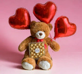 A Jumbo brown teddy bear with a box of chocolates and 3 heart shaped red balloons on a pink backdrop.