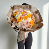 A bouquet of yellow, peach and white roses, wrapped with kraft paper, hold by a person, against a white backdrop.