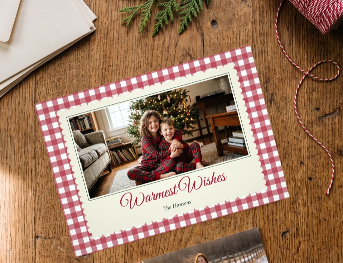 Two family holiday photo cards, Warmest Wishes in red gingham and JOY, on a wooden table with envelopes and twine
