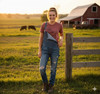 Small Town Girl Graphic Tee Tshirt, Clay — Smiling woman in overalls standing by a wooden fence at sunset