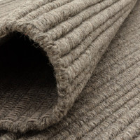Close-up of a rolled greige wool rug demonstrating its flexibility, thickness, and natural yarn weave.