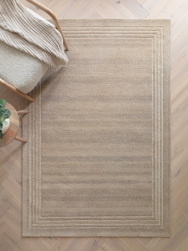Wide lifestyle shot of a spacious, bright interior styled with a large sand beige wool area rug on light oak hardwood flooring.