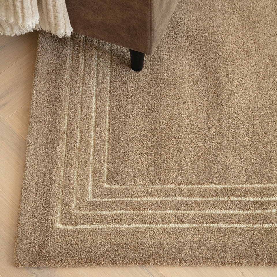 Detailed corner view of a brown tufted wool rug with a triple cream border, styled on a light oak herringbone floor under an armchair leg.