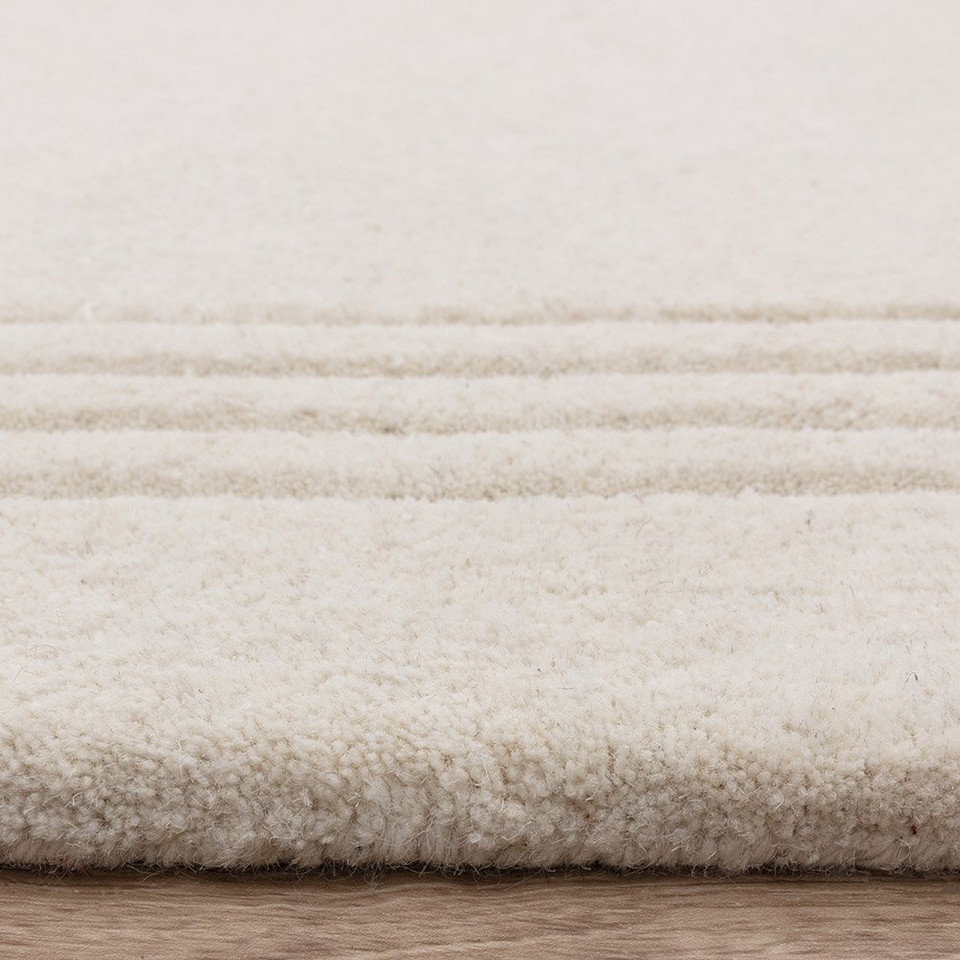 Close-up profile of a plush ivory wool rug featuring a three-line charcoal grey accent border on a light wood floor.