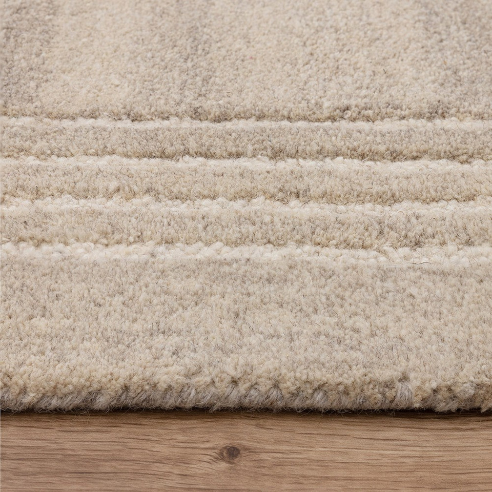 Close-up profile of the Orlo sand beige wool rug showing its thick plush texture and dark grey triple pinstripe border on a light wood floor.