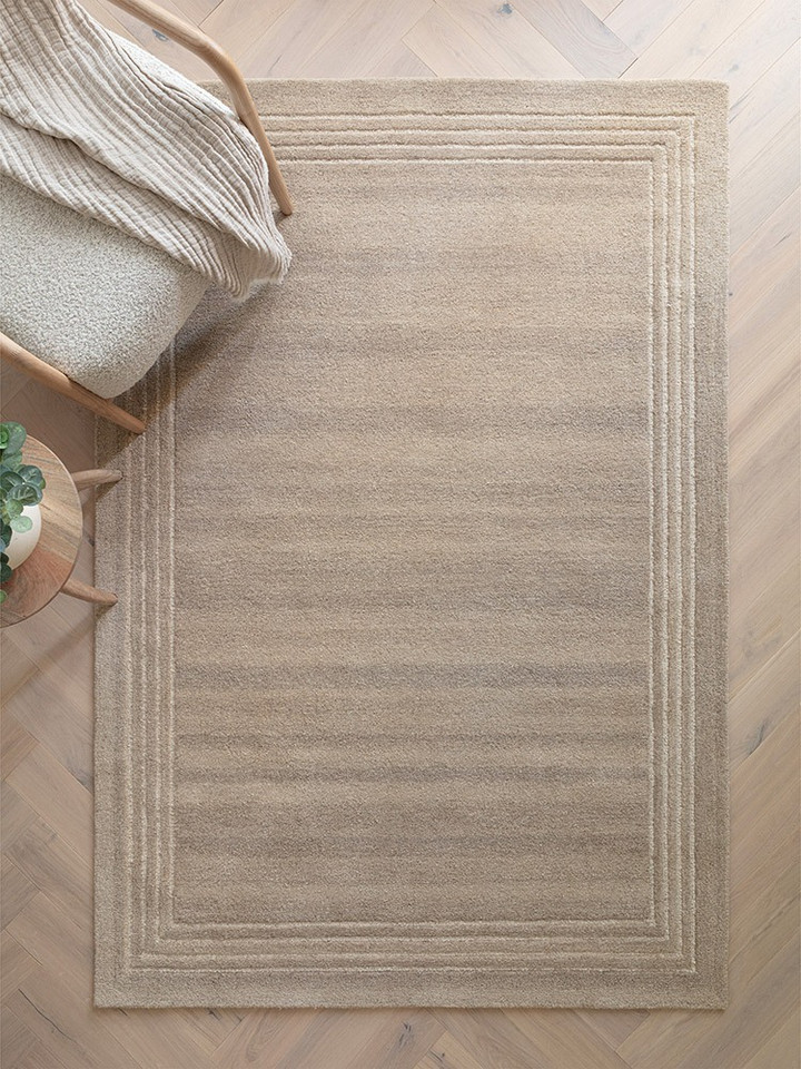 Wide lifestyle shot of a spacious, bright interior styled with a large sand beige wool area rug on light oak hardwood flooring.