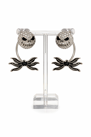 Features skull stud earrings with a separate spider bow backing designed to sit behind the ear, creating a layered, dimensional look. Images include clean white background product shots for detail, a stand display for structure, an on-ear lifestyle photo to show fit and placement, and a dark, moody flat lay to highlight the gothic aesthetic and sparkle.