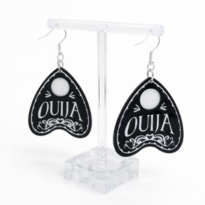 Collection of Ouija planchette acrylic earrings displayed in multiple design variations on a clean background. Each pair features unique lettering styles, symbols, and finishes while maintaining a cohesive dark, witchy aesthetic. The assortment highlights different options within the same occult-inspired theme.
